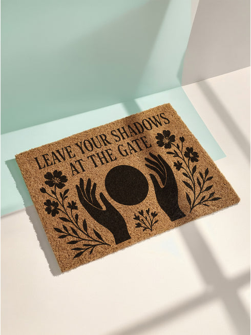 A rectangular, natural brown coir welcome mat with a textured surface, featuring an intricate, black witchy design intended for spiritual protection. The celestial and metaphysical pattern showcases a central manifestation spell circle with sacred geometry, radiating lines, and is accented with stars and crescent moons at the corners, creating a unique piece of entryway decor.
